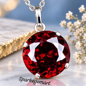 May include: A round, deep red faceted gemstone pendant set in a silver-toned metal prong setting. The pendant hangs from a silver-toned chain. The text "Sparkegemart" is visible at the bottom of the image. The stone features a brilliant cut.