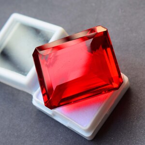 May include: A large, faceted, rectangular red gemstone, possibly a ruby or garnet, is displayed in a white plastic case. The gem has sharp edges and a brilliant, reflective surface. The case is open, revealing the gem's vibrant color.