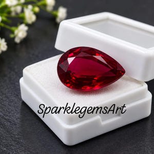 Könnte beinhalten: Ein birnenförmiger, tiefroter facettierter Edelstein in einer kleinen, offenen weißen Kunststoff-Präsentationsbox. Der Text SparklegemsArt ist in Schwarz auf der Vorderseite der Box aufgedruckt. Der Edelstein weist einen Brillantschliff auf.