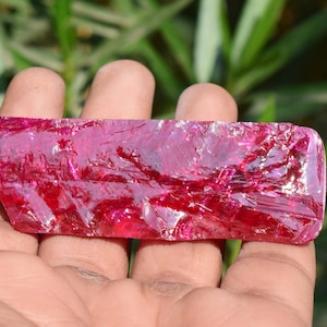 May include: A rough-cut, rectangular gemstone with a deep magenta hue. The translucent stone has a fractured, crystalline appearance, catching light and reflecting a range of pink and red tones. The gem is held in an open hand against a blurred green background.