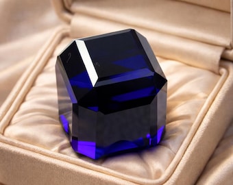May include: A faceted, dark blue crystal ring, displayed in a beige jewelry box. The ring has a geometric design with multiple sides, reflecting light. The box is lined with a soft, satin-like material, enhancing the ring's presentation.