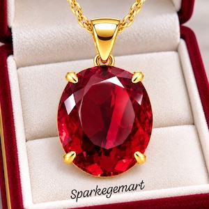 May include: A gold pendant necklace featuring a large, oval-cut, deep red gemstone held by four prongs on a gold chain. The jewelry is displayed in a red velvet box with the text Sparkegemart printed in black on the interior lining.