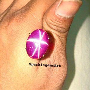 Peut inclure: Une pierre précieuse rubis étoilé de forme ovale et de couleur magenta. La gemme présente un motif en étoile à six rayons. Le texte "SparklegemsArt" est visible en bas de l'image.