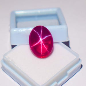 Puede incluir: Una gema ovalada de color rojo rubí vibrante con un patrón de líneas blancas en forma de estrella. La gema se exhibe en una caja de joyería abierta, azul claro y blanca, sobre un fondo blanco. El efecto de estrella es prominente.
