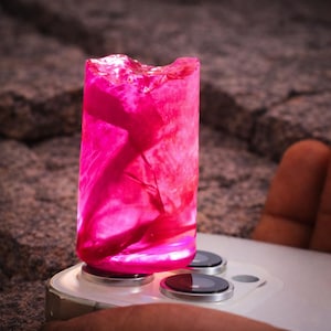 May include: A vibrant, translucent pink cylindrical object, possibly a decorative light or art piece, is illuminated. The object is placed on a smartphone, with the phone's camera lenses visible. The background is a textured, rocky surface.
