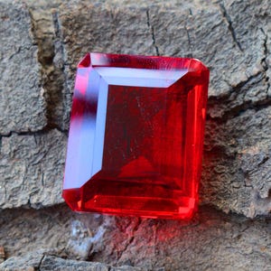 May include: A rectangular-cut, deep red gemstone with multiple facets. The gem has a polished surface and sharp edges. The stone is set against a textured, brown background, highlighting its vibrant colour and clarity.