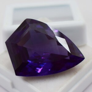 May include: A deep purple, faceted gemstone with a triangular, shield-like shape. The stone features multiple geometric cuts that create sharp edges and reflective surfaces, showcasing a rich, saturated violet colour throughout the entire piece.