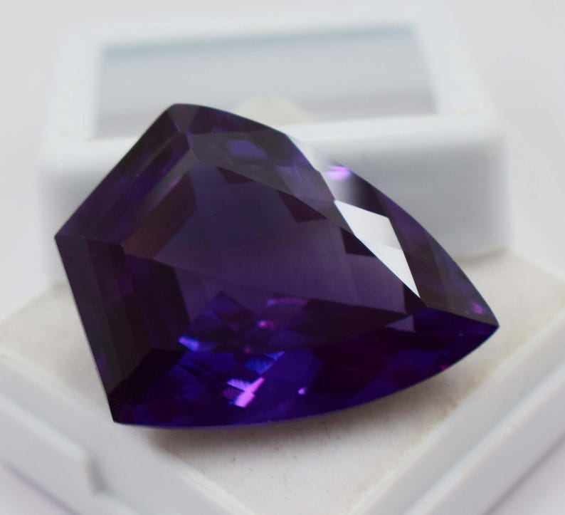 May include: A deep purple, faceted gemstone with a triangular, shield-like shape. The stone features multiple geometric cuts that create sharp edges and reflective surfaces, showcasing a rich, saturated violet colour throughout the entire piece.