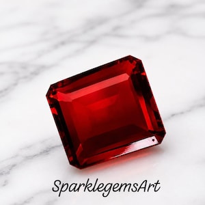 May include: A square-cut, deep red gemstone with multifaceted surfaces that reflect light. The stone features a brilliant, transparent finish and is positioned above the black text SparklegemsArt, which is written in a cursive script font.