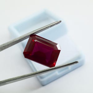 May include: A faceted, rectangular ruby gemstone held by tweezers. The gem is a deep, rich red color and has a polished surface. The tweezers are silver-colored, and the background includes a light blue plastic case.