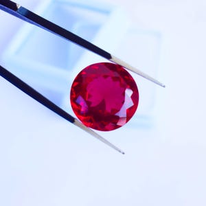 May include: A vibrant, round, deep red gemstone is held by tweezers against a light blue backdrop. The gem's facets reflect light, creating a sparkling effect. The tweezers are black with silver tips. The gem is likely a ruby.
