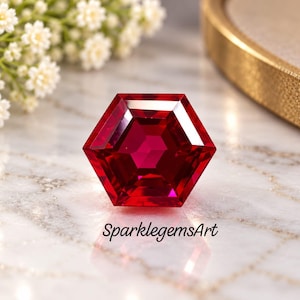 May include: A vibrant, deep red gemstone cut into a precise hexagonal shape with multiple facets that reflect light. The stone is positioned on a white marble surface, and the text SparklegemsArt is printed in black below the gemstone.