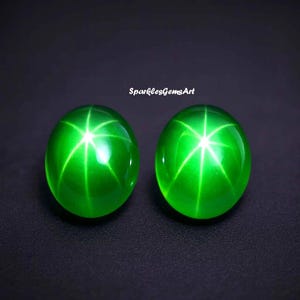 May include: Two oval-shaped, vibrant green gemstones with a six-pointed star effect. The gems are highly polished, reflecting light and set against a dark background. The text "SparklesGemsArt" is visible.