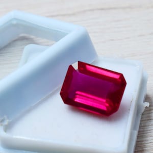Può includere: Una vibrante gemma rosso rubino, taglio rettangolare, è posta in un astuccio di plastica azzurra aperto. Le sfaccettature della gemma riflettono la luce, mostrando il suo colore e la sua chiarezza. L'astuccio offre uno sfondo pulito e contrastante.