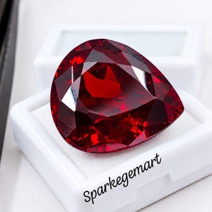 May include: A pear-shaped, deep red faceted gemstone resting in a white display box. The box features the black text Sparkegemart printed on the front. A pair of black metal tweezers is positioned to the left of the gemstone.