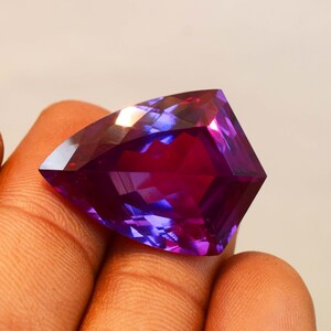 May include: A faceted trillion-cut gemstone displaying a vibrant gradient of deep purple and magenta hues. The stone features a triangular shape with sharp, clean edges and a polished surface that reflects light, showcasing a translucent colour transition.