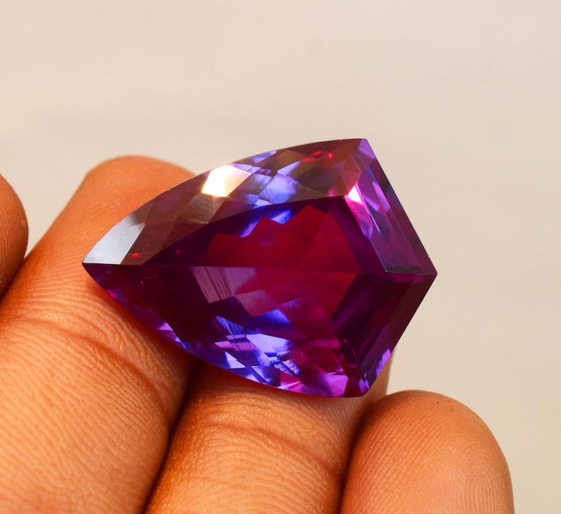 May include: A faceted trillion-cut gemstone displaying a vibrant gradient of deep purple and magenta hues. The stone features a triangular shape with sharp, clean edges and a polished surface that reflects light, showcasing a translucent colour transition.