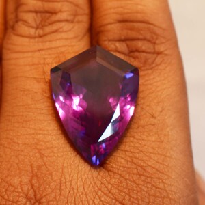 May include: A shield-shaped faceted gemstone displaying a deep purple and violet colour gradient. The stone features a pointed bottom and a flat top edge with multiple triangular facets that reflect light, creating a vibrant, translucent appearance.