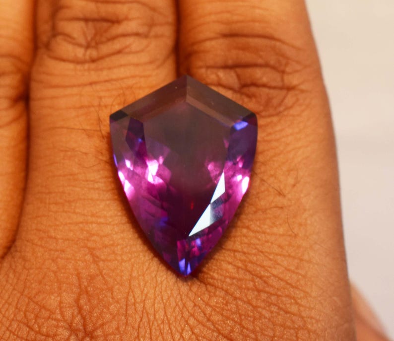 May include: A shield-shaped faceted gemstone displaying a deep purple and violet colour gradient. The stone features a pointed bottom and a flat top edge with multiple triangular facets that reflect light, creating a vibrant, translucent appearance.