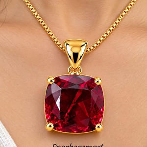 May include: A gold necklace featuring a large, square-cut, deep red gemstone pendant held by four prongs. The pendant is suspended from a gold box chain. The text Sparkegemart is printed in black below the pendant on the white fabric background.