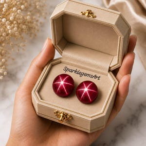 Könnte beinhalten: Zwei runde, tiefrote Cabochon-Edelsteine mit einem ausgeprägten sechsstrahligen Sterneffekt, präsentiert in einer beigen, achteckigen Schmuckschatulle mit goldenem Verschluss. Der Text SparklegemsArt ist schwarz auf das Innenfutter gedruckt.