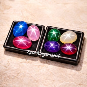 May include: Two black trays each hold six oval gemstones in various colours, including blue, pink, green, yellow, purple, and red. Each gem has a star-like pattern. The text "SparklegemsArt" is visible.