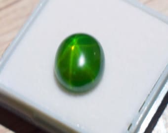 Puede incluir: Una gema verde vibrante con un patrón en forma de estrella, colocada sobre un fondo blanco dentro de un estuche gris. La superficie pulida refleja la luz, mostrando la claridad y el color de la gema. Este cabujón es un ejemplo llamativo de una piedra preciosa natural.