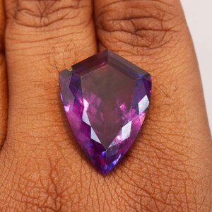 May include: A shield-shaped faceted gemstone displaying a vibrant gradient of deep purple and magenta hues. The stone features a polished surface with multiple geometric facets that reflect light, creating a brilliant and translucent appearance.