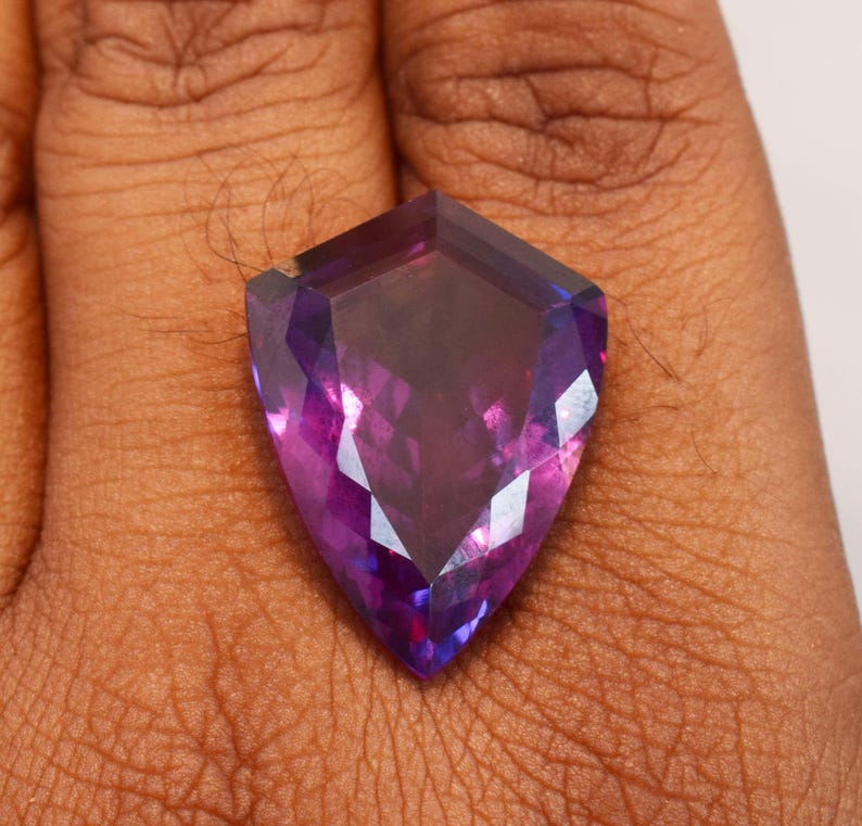 May include: A shield-shaped faceted gemstone displaying a vibrant gradient of deep purple and magenta hues. The stone features a polished surface with multiple geometric facets that reflect light, creating a brilliant and translucent appearance.