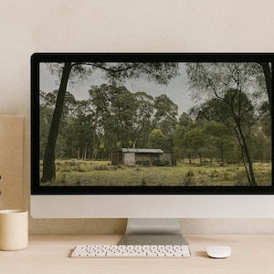 Może przedstawiać: Komputer wyświetla obraz małej chaty w lesie. Klawiatura, mysz, beżowy kubek, szklany wazon z zielenią i brązowy papier znajdują się na drewnianym biurku.