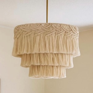 Puede incluir: Una lámpara de araña de tres niveles con una pantalla de macramé beige y flecos. La lámpara tiene un vástago y un soporte de techo dorados. La lámpara de araña está suspendida del techo, un elemento de iluminación decorativo.