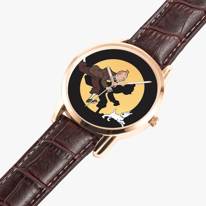 Op de afbeelding: Goudkleurig polshorloge met een bruine leren band. De wijzerplaat toont een cartoonillustratie van een man en een hond tegen een gele en zwarte achtergrond. De wijzers van het horloge zijn zilver.