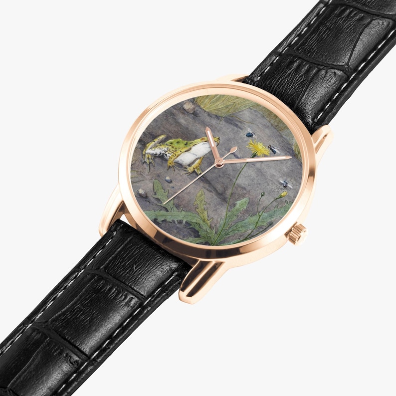 May include: A rose gold wristwatch with a black leather band. The watch face features a detailed illustration of a frog, plants, and insects. The watch has a classic design with a round face and a textured leather strap.