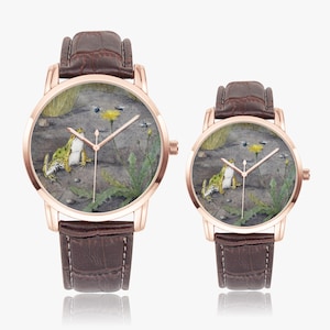 May include: Two rose gold watches with brown leather straps. The watch faces feature a detailed painting of a frog, a yellow flower, and insects. The watch hands are gold. The watches are displayed on a white background.