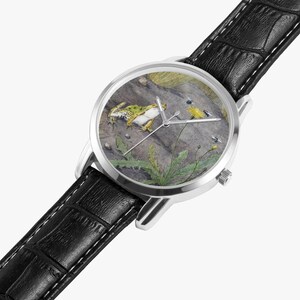 May include: A silver wristwatch with a black leather band. The watch face features a detailed illustration of a frog, insects, and plants on a rock. The watch has silver hands and a silver case.