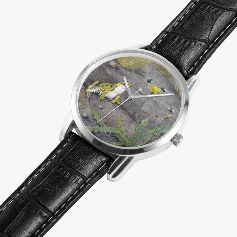 May include: A silver wristwatch with a black leather band. The watch face features a detailed illustration of a frog, insects, and plants on a rock. The watch has silver hands and a silver case.