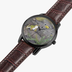 May include: A black wristwatch with a brown leather band. The watch face features a detailed illustration of a frog, insects, and plants. The band is approximately 0.75 inches wide and has a textured, crocodile-like pattern.