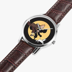 Op de afbeelding: Een polshorloge met een bruine leren band en een zilverkleurige kast. De wijzerplaat toont een cartoonillustratie van een man, een hond en een gele achtergrond. De wijzers van het horloge zijn zilver.