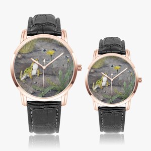 May include: Two rose gold watches with black leather straps. The watch faces feature a detailed illustration of a frog, a yellow flower, and insects in a naturalistic setting. The watches are displayed on a white surface.