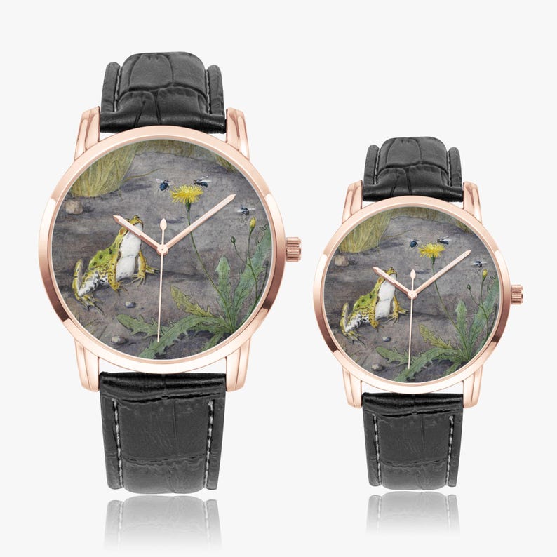 May include: Two rose gold watches with black leather straps. The watch faces feature a detailed illustration of a frog, a yellow flower, and insects in a naturalistic setting. The watches are displayed on a white surface.