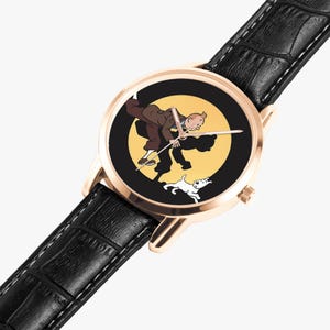 Op de afbeelding: Een goudkleurig polshorloge met een zwarte leren band. De wijzerplaat toont een cartoonillustratie van een man, een hond en een gele achtergrond. De wijzers van het horloge zijn zilver.
