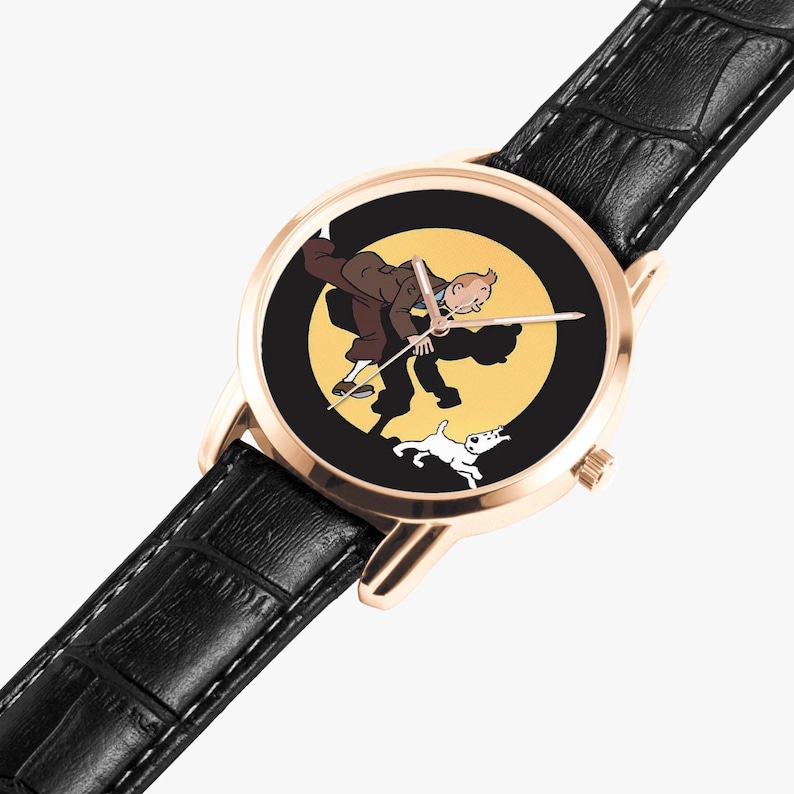 Op de afbeelding: Een goudkleurig polshorloge met een zwarte leren band. De wijzerplaat toont een cartoonillustratie van een man, een hond en een gele achtergrond. De wijzers van het horloge zijn zilver.