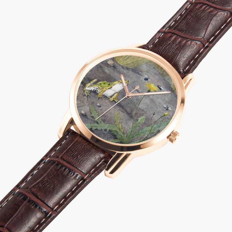 May include: A gold-tone wristwatch with a brown leather band. The watch face features a detailed illustration of a green frog on a rock with plants and insects. The watch hands are gold-tone.