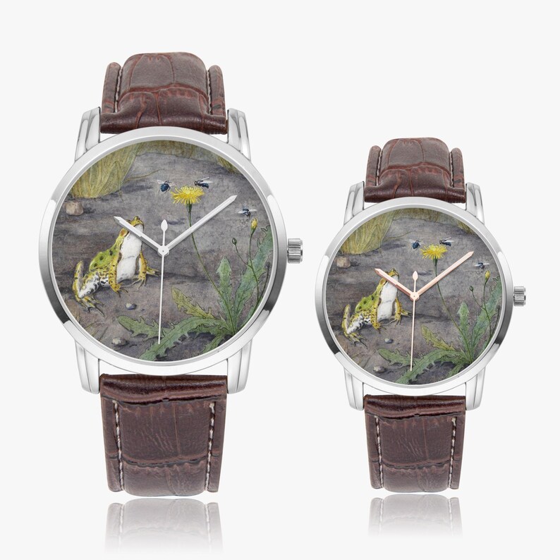 May include: Two silver-tone wristwatches with brown leather bands. The watch faces feature a detailed illustration of a frog, a dandelion, and insects. The watch hands are silver and rose gold. The watches are displayed on a white background.