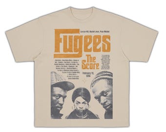 May include: Cream-colored t-shirt featuring a graphic design with the word "Fugees" in orange block letters. The design includes the text "The Score" and the date "February 13, 1996", along with portraits of three people.