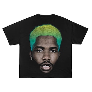Op de afbeelding: Zwart T-shirt met een portret van een persoon met groen en geel haar. Het gezicht is een close-up, met een pixelachtig effect. Het T-shirt heeft een ronde hals, een veel voorkomende stijl voor vrijetijdskleding.
