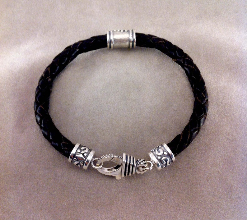 May include: A black leather bracelet with silver-toned clasps and decorative accents. The bracelet is braided and has a simple design.