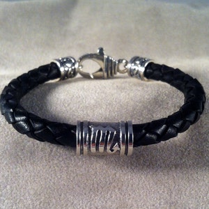 May include: A black leather bracelet with a silver clasp and a silver bead with a design.