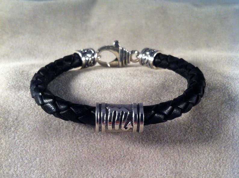 May include: A black leather bracelet with a silver clasp and a silver bead with a design.
