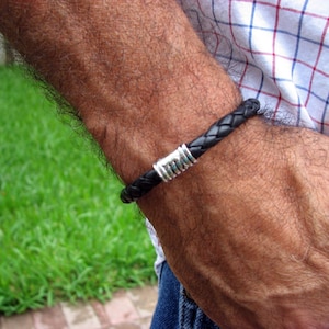 May include: A black leather bracelet with a silver metal clasp. The bracelet is worn on a person's wrist.